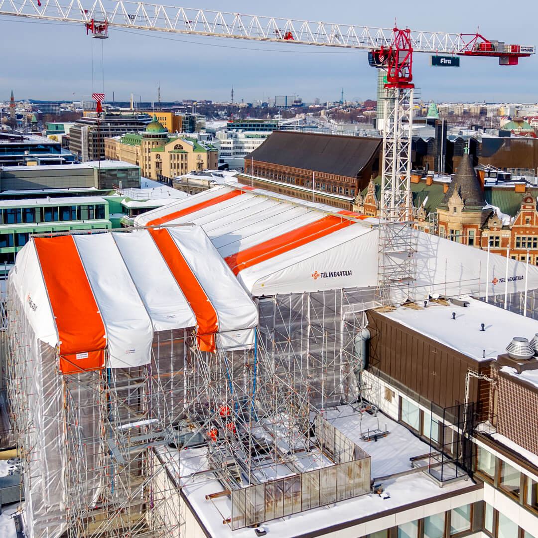 Vy över taken med vita och orange väderskyddstak från Telinekataja som täcker ställningarna, tornkranar ovanför och det snötäckta Helsingfors centrum i bakgrunden.