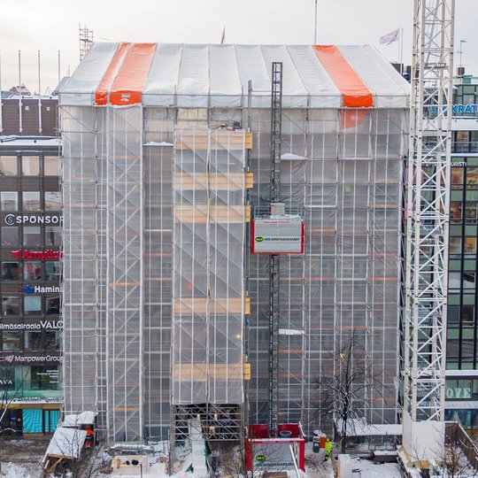 Kontorsbyggnad vid Mannerheimvägen i Helsingfors helt inbyggd i ställningar och genomskinlig skyddsplast, med ett vitt och orange väderskyddstak överst och en röd bygghiss längs fasaden en snöig vinterdag.