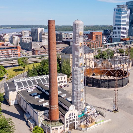 Flygbild över Suvilahti i Helsingfors med en hög skorsten inklädd i ställningar och väderskydd under renovering, intill den historiska tegelskorstenen och gasklockorna.