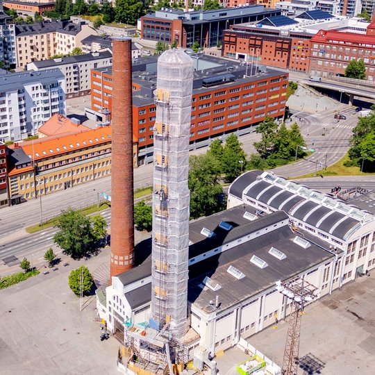 Flygbild över två skorstenar som reser sig över industribyggnaderna i Suvilahti i Helsingfors, den ena inklädd i ställningar för renovering.