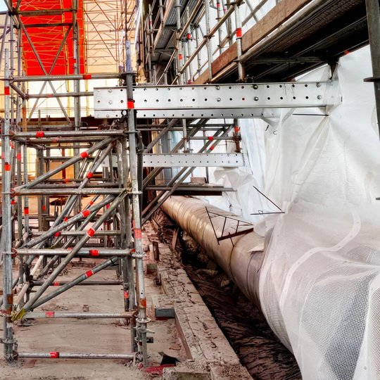 Inuti ställningskonstruktionen: galvaniserade ställningsrör och stålbalkar vid sidan av skyddsplast som täcker en öppnad takkonstruktion under renovering.